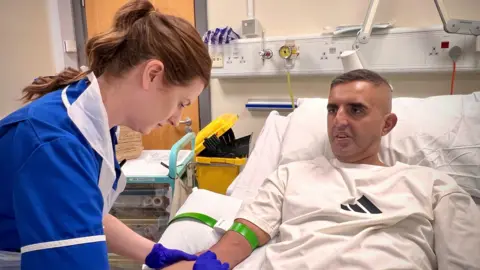 A man with short dark hair is sitting in a hospital bed and is having some blood taken from his arm by a nurse.