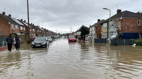 PA Media A flooded residential street with two people wading along the road in water up to the bumpers of cars