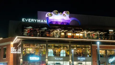 Andrew Scott A large purple five-eyed inflatable monster next to the 'everyman' sign. Below is an outdoor bar, which is above a selection of shops.