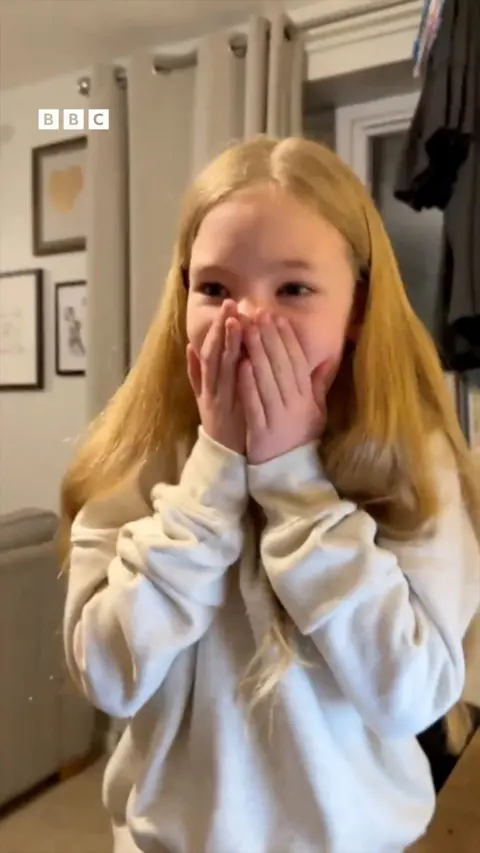 A young girl with blonde hair, wearing a white jumper.