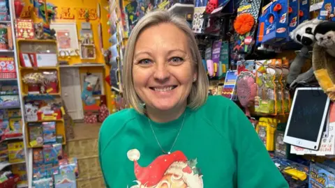 Charlotte Crosser in a green jumper with the top of Father Christmas, including his hat visible. She is stood in a shop with toys and gifts behind her.