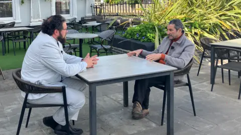 Two men facing each other, seated at an outdoor table in a beer garden. The man on the left has bushy black hair, a beard and wears a pale suit and black boots. The man on the right has slicked-back, dark grey hair and wears a grey jacket over orange jumper and black trousers.