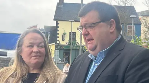 Donna-Marie McGillion has long blond/brown hair and is wearing a tan coloured coat and black top. She is looking at her husband Garry who is speaking. Garry has short dark hair and black-framed glasses and is wearing a black suit jacket and blue shirt. A pub is in the background of the picture.