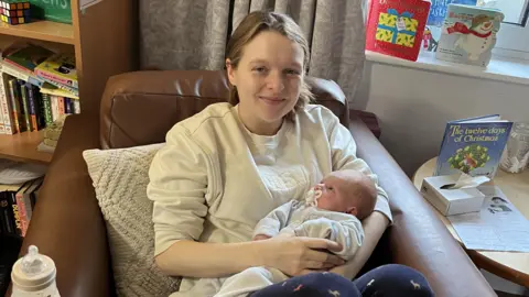 Family handout A woman smiles at the camera while holding a baby. She is sat in a brown chair.