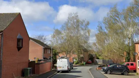 Google A street view image of a road with houses on each side and cars parked on the road. The homes are made of red brick and there is a white van on the road nearest the camera. A couple of trees are next to the homes.