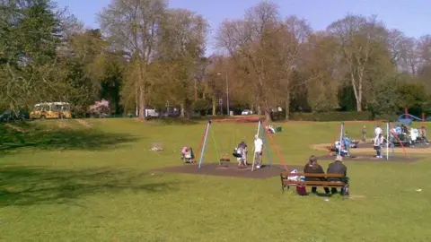 Burgess Von Thunen/BBC Large grass park with trees in the background. There is a playground in the foreground with swings and a toy train. There are children playing on the equipment and some adults are sitting on a bench. There is a yellow and white ice cream van in the background.