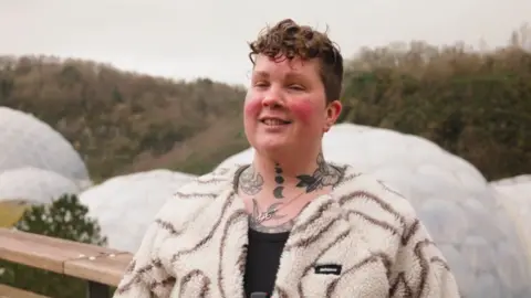 Eden Project A woman wearing a white fleece and black t-shirt, with short curly brown hair, standing in front of the Eden Project biomes