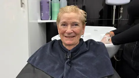 Ben Schofield/BBC Pat Larham, having her hair washed, sitting in a hairdresser's chair, with a blue towel round her neck, with blonde short hair, a sink behind her and earrings in both ears. She is smiling at the camera. 
