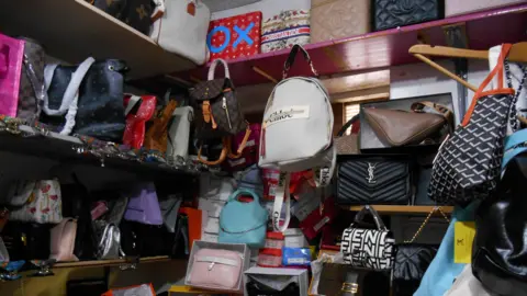Shelves packed with a large collection of fake designer handbags in various colours and patterns. They are in a small storage room.