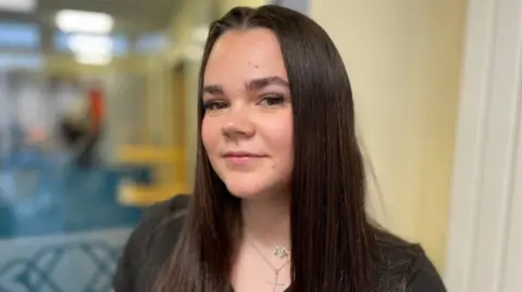 Daisy has long straight brown hair and dark eyes. She is wearing a black t-shirt and has two silver necklaces around her neck. 