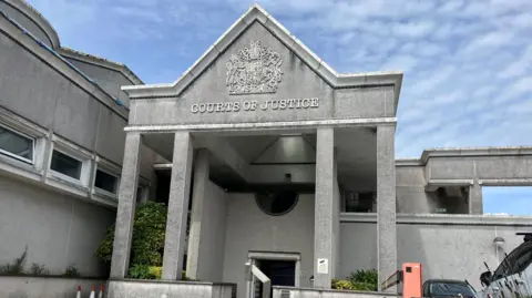 The front entrance of Truro Crown Court. It has four granite pillars and the lettering: COURTS OF JUSTICE with an elaborate shield above. There is a car park to the right and the ski is blue with light cloud.
