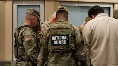 A photograph of a number of National Guard members standing together. At least 5 men can be seen in the image - one of whom appears to be in civillian clothing wearing a cream-coloured shirt. The rest of the men are in uniform and the words 'National Guard' can be seen on the back of their uniform.