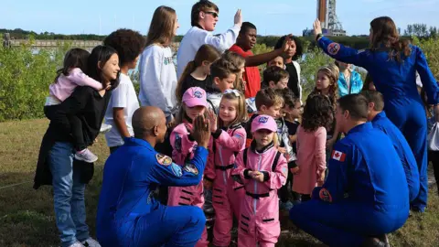 Joe Raedle/Getty Images On a grassy patch near water, three Artemis II astronauts in bright blue flight suits crouch down to talk with a group of young children. The children in the centre wear miniature pink spacesuits and caps, facing the astronauts and giving them high‑fives. Other children and parents cluster around them, some holding toddlers, forming a loose semicircle. In the distance, partly blurred, a tall rocket and launch tower rise above the trees. The mood is warm and playful, with astronauts and families smiling and interacting at eye level, turning a serious mission into a friendly, down‑to‑earth moment.