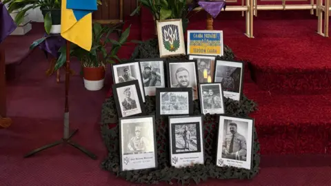 Ewhen Chymera Several black-and-white pictures are displayed on red steps commemorating people who have died in the conflict in Ukraine. The bottom of a Ukrainian flag can be seen on the left of the picture.