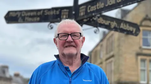 A grey-haired man with glasses stands beneath sign-posts to historic attractions, wearing a blue fleece