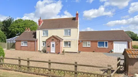 A large, white-rendered, detached home, including single storey extensions on each side. In front of the property is a large gravel driveway and an outbuilding with a low wooden fence surround the perimeter.  