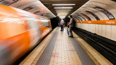A subway carriage pulling into St George's Cross station blurred. People stand on the platform.