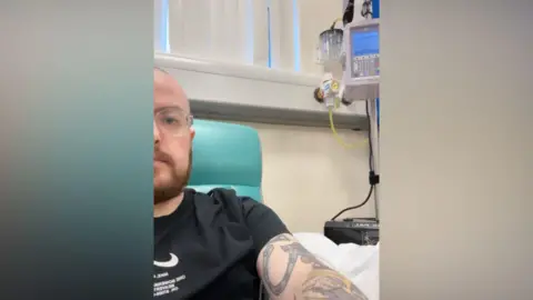 Robbie Meakin A man with glasses, a short beard and tattoos on his arm sits in a green hospital chair wearing a black T-shirt. Medical equipment including a monitor and tubing is visible on the wall beside him. Daylight comes through vertical blinds in the background.