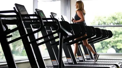 A stock image of a woman running on a treadmill. She is in a gym with floor to ceiling windows in front of her and next to her. She is running on one of six treadmills lined up and the others are empty.