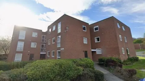 Google Streetview of a block of three-storey red brick flat buildings, that have lots of windows and flat roofs. The linked buildings with multiple entrances are at angles to each other, each main block being roughly square shaped.