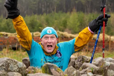 Chris Daniel A man at the bottom of Pen y Fan. There are rocks around him. The man has a blue t-shirt and under a yellow long sleeved shirt with a blue hat, black sunglasses and black shorts. His arms are outstretched like his is cheering. 