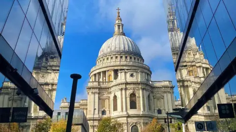 St Paul's Cathedral, a white stone building surrounding by glass buildings which share its reflection. 