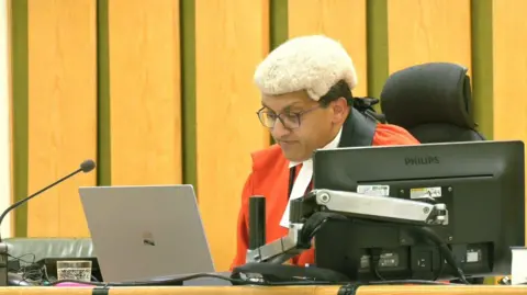 PA Media The judge, wearing red robes and a wig, reads from a laptop at the bench. He is an asian man and is wearing glasses. 