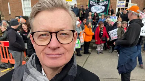 Lucinda Adam/BBC Neil Blanchard has short blond hair and wears glasses. He stand with the demonstration behind him wearing a black coat and a patterned grey scarf. 