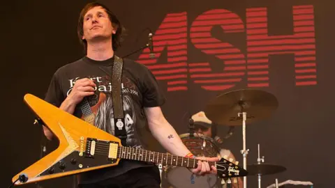 Tim Wheeler of Ash wears a dark Kurt Cobain T-shirt and a pale wood guitar. He is on stage with a drummer behind him and the word ASH in red on a black screen behind him