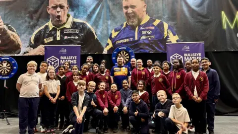 BBC A group of school children standing infront of darts boards and some adults are also in the photo. They are smiling and some have their thumbs up. Their school uniform is maroon with a yellow crest. 