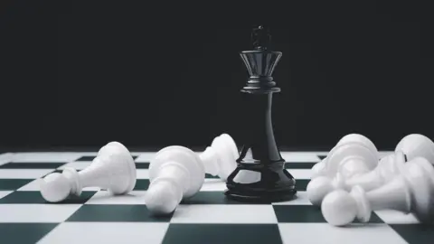 Getty Images On a black and white chess board, a black king chess piece stands upright. It is surrounded by toppled white pawns.