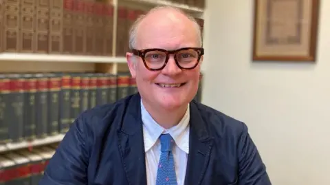 Gary Rycroft Gary Rycroft wearing thick tortoise shell rimmed glasses and a suit with a baby blue tie in an office with legal books in the background