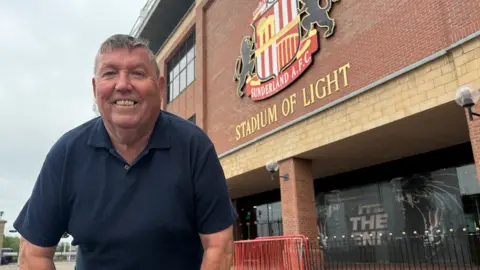Peter Farrer stands wearing a navy blue polo shirt, and smiling to the camera, in front of a building with the words Stadium of Light printed in gold and the club's crest above. Towards the bottom of the picture, a graphic has been added to the windows which says ''Til The End'.
