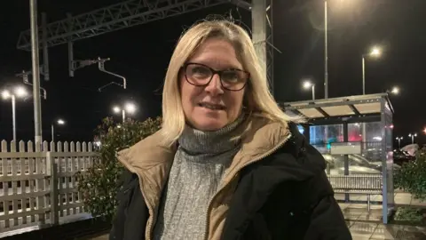 Alex Pope/BBC Woman with blond hair and glasses at a railway station