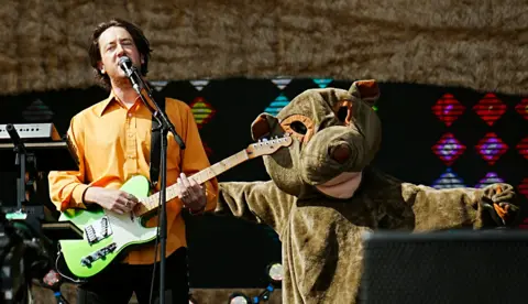 The Wombats singer, wearing an orange shirt, sings and plays a guitar in front of a person dressed in a wombat suit