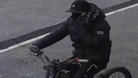 Merseyside Police A still image of a man riding a red handled electric bike. He is dressed all in black with a bubble style coat, tracksuit bottoms, gloves, a baseball cap and a  covering over his lower face. His left hand is holding a mobile phone to his ear while his right hand is gripping the handle-bars of the bike. 