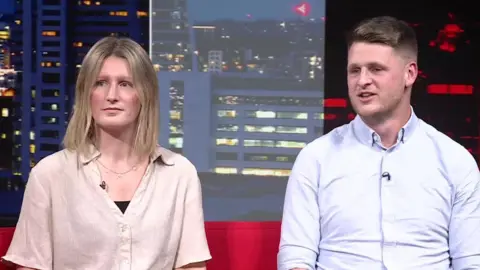 Laura and Ben are seated on a bright red couch in a studio setting. Laura is wearing a light beige short-sleeved shirt over a black inner layer and has a necklace. Ben is wearing a light blue button-up shirt with rolled-up sleeves. Behind them is a large backdrop showing a cityscape at night with illuminated buildings.