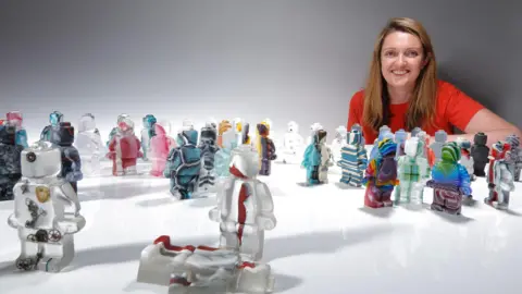 A woman crouching behind a table, smiling at the camera. On the table are glass figurines of Lego men