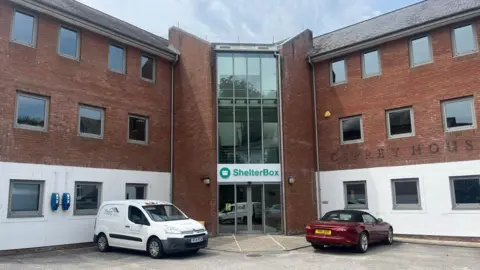 The outside of the new ShelterBox office on Malpas Road, Truro. The company logo is above glass doors. There are two vehicles parked outside.