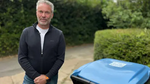 Martin Giles/BBC A man is standing on a drive-way with a hedge in the background. In the Foreground there is a bin with a bright blue lid. The mans is wearing denim jeans and a blue jumper. 