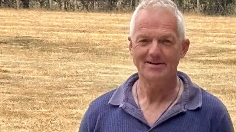 Luke Deal/BBC A man stands in a field and smiles at the camera. He has short grey hair and wears a blue polo shirt.