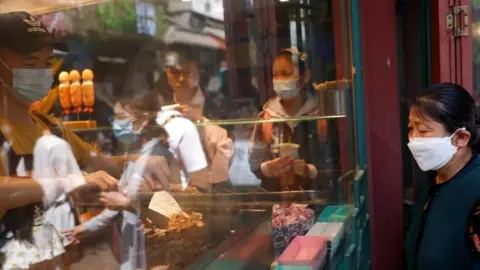 Reuters A woman wearing a face mask looks at a man cooking inside a restaurant in Beijing