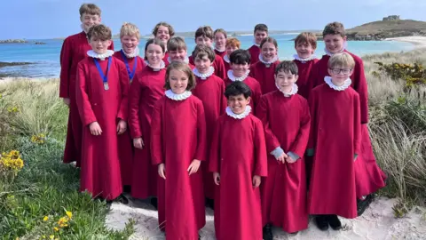 A group of boys and girls wearing choir surplices stand on a beach on the Isles of Scilly 