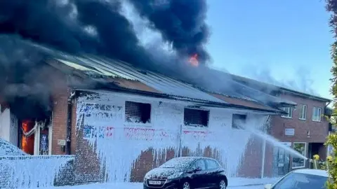 A red-brick commercial building is on fire with large plumes of black smoke coming from the roof and flames in the doorway. There is a jet of foam spraying into one of three windows and the front of the building is covered with more foam. There is a car parked outside the building.