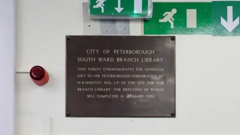 Friends of Woodston Library A brown plaque put up on a wall says: "CITY OF PETERBOROUGH SOUTH WARD BRANCH LIBRARY. This tablet commemorates the generous gift to the Peterborough corporation by HB Hartley ESQ JP of the site for this branch library, the erection of which was completed in January 1951".