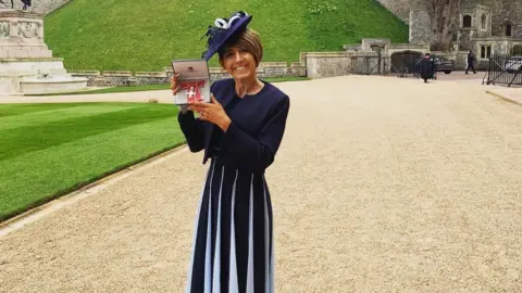 Natalie Queiroz A woman with short brown hair is wearing a navy blue dress with white slits. She is wearing a navy and white hat. She is holding a red MBE medal in a box, and is standing in front of a green garden