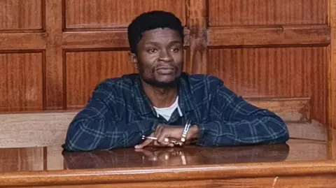 Webster Ochora Elijah in a checked shirt, behind the dock in court