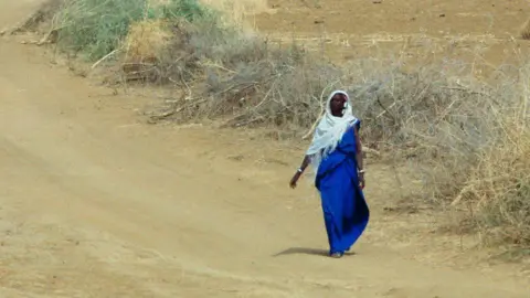Getty Images A woman wearing a blue sari and white head piece. She is walking along a dry sand path with dry scrub to her right.