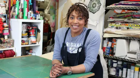 Jacqui Joseph, in a blue top and black apron, stands in an art room with paint on shelves behind her.
