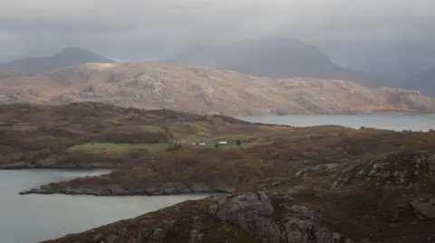 Getty Images A landscape of lochs and hilly land with rocky outcrops and heather covered slopes. A small white-walled house sits amongst this landscape, and there are mountains behind mist and low cloud in the distance.